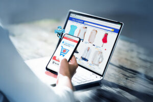 Dłoń trzymająca telefon z otwartą stroną koszyka sklepu internetowego, w tle laptop z otwartą stroną sklepu internetowego